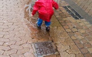 Two young children, both wearing bright red raincoats and hoods, play on a wet, brick-paved pavement. One child squats down to touch a puddle, while the other stands nearby. The ground is wet with rain, and puddles reflect the grey sky.