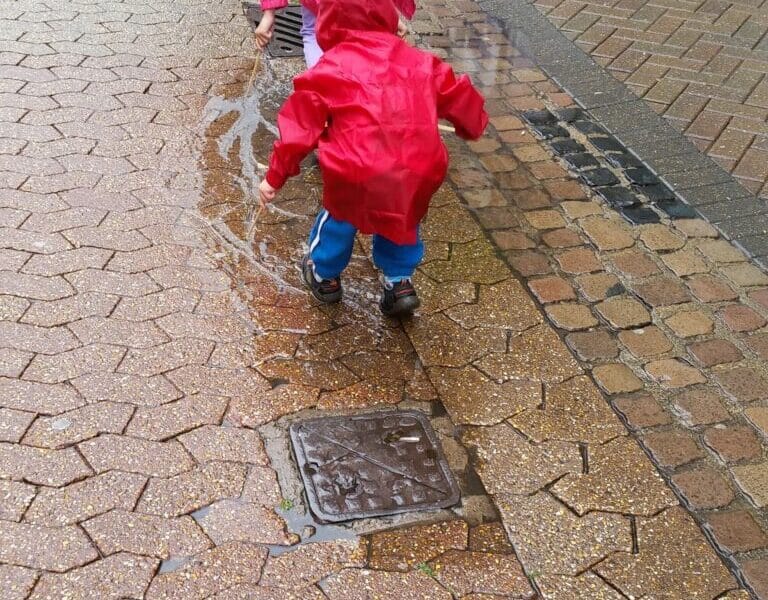 Two young children, both wearing bright red raincoats and hoods, play on a wet, brick-paved pavement. One child squats down to touch a puddle, while the other stands nearby. The ground is wet with rain, and puddles reflect the grey sky.