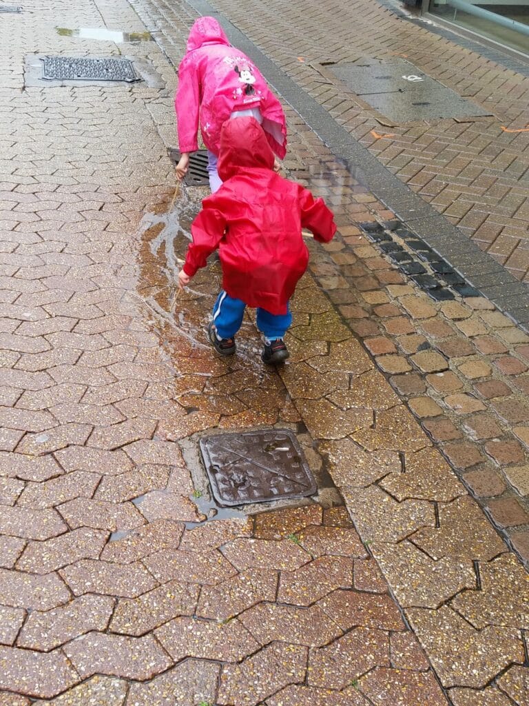 Two children in bright red raincoats and hoods walk on a wet, brick-paved pavement. They lean forward, watching water flow along the edge of the pavement, with puddles reflecting the rainy weather. One child wears blue trousers, the other striped leggings.