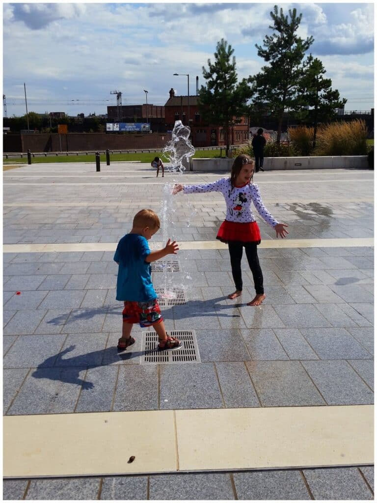 Two children play in a square; a girl in a white jumper and red skirt stands smiling near a water fountain, arms open, while a boy in blue shorts and a red shirt steps on a water grate. Trees, grass, and buildings are in the sunny background.