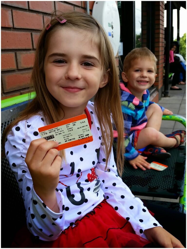 A young girl with light brown hair, wearing a white polka dot shirt and red skirt, smiles while holding a train ticket. A smiling younger boy in a striped shirt and shorts sits beside her on a bench, with a brick wall and people blurred in the background.