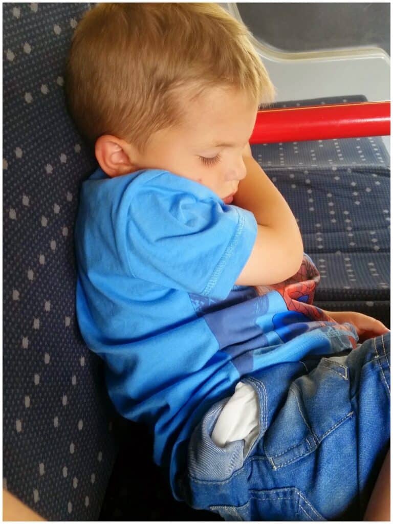 A young boy with blond hair, wearing a blue T-shirt and denim shorts, is sleeping while sitting on a dark blue patterned seat. His head rests on his bent arm, eyes closed, and he is leaning sideways against the back of the seat.