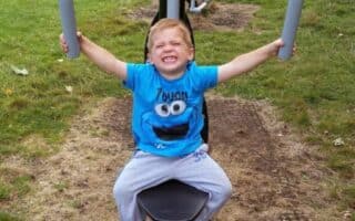 A young boy with blond hair, wearing a blue Cookie Monster T-shirt and grey trousers, sits smiling on an outdoor exercise machine in a grassy park. He holds onto the handles above his head, with trees and another person on exercise equipment behind him.
