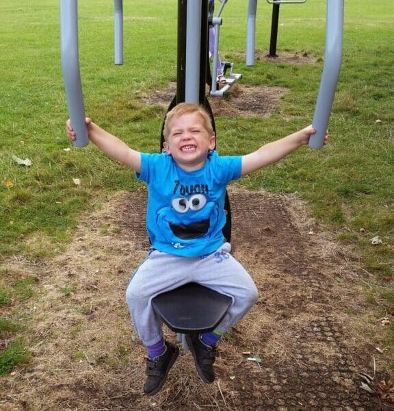 A young boy with blond hair, wearing a blue Cookie Monster T-shirt and grey trousers, sits smiling on an outdoor exercise machine in a grassy park. He holds onto the handles above his head, with trees and another person on exercise equipment behind him.