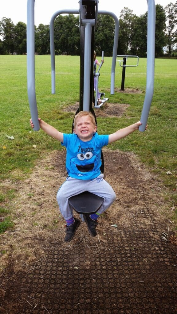 A young boy in a blue Cookie Monster shirt and grey tracksuit bottoms smiles widely whilst sitting on outdoor gym equipment in a grassy park; his arms are raised holding the handles. Green trees and more equipment are visible in the background.
