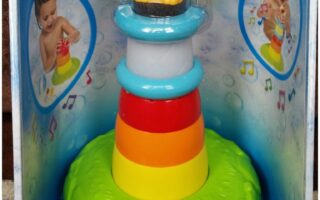 A colourful TOMY Stack & Play Lighthouse toy is shown in its packaging. The toy has stackable, rainbow-coloured rings and a yellow character wearing goggles and a raincoat on top. Images of children playing with the toy are featured on the box.