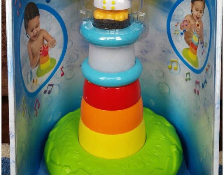 A colourful TOMY Stack & Play Lighthouse toy is shown in its packaging. The toy has stackable, rainbow-coloured rings and a yellow character wearing goggles and a raincoat on top. Images of children playing with the toy are featured on the box.