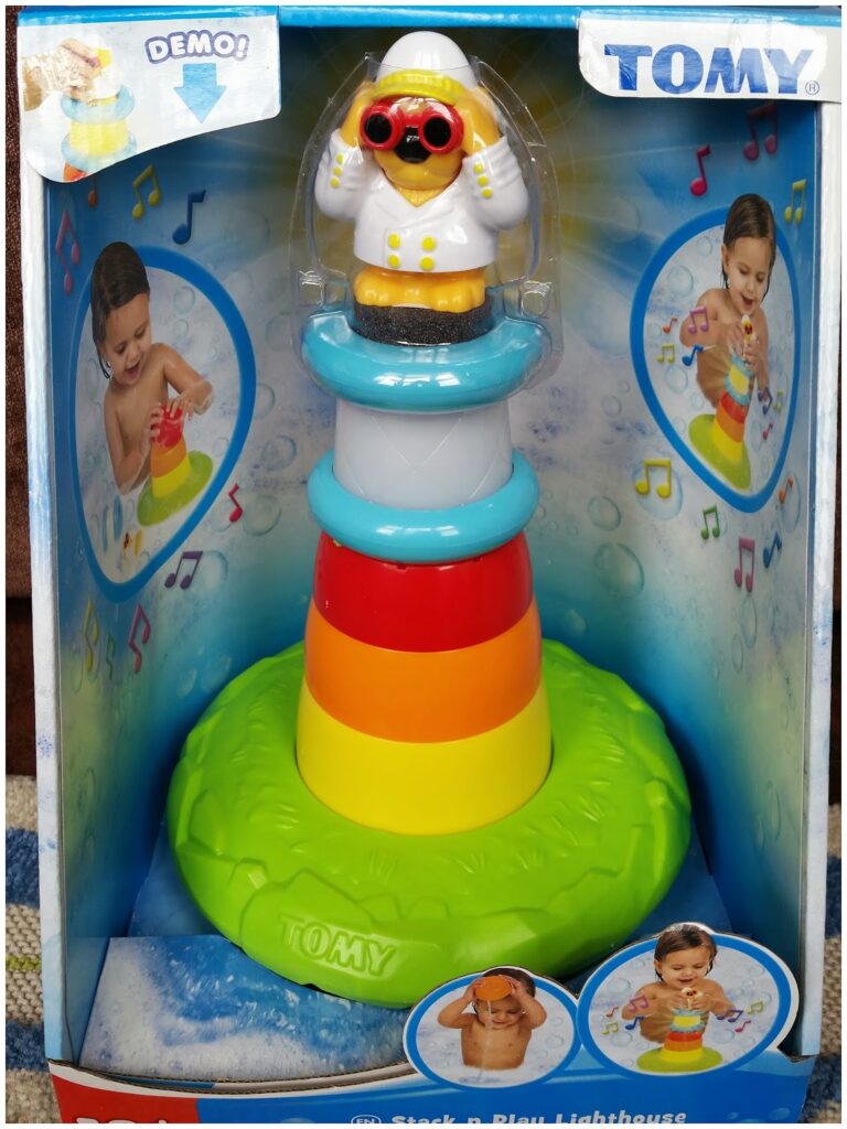 A boxed TOMY toy called "Stack & Play Lighthouse" features a colourful lighthouse with red, orange, yellow, blue, and white rings and a bird in a raincoat and goggles on top. Two children are shown playing with it in water on the packaging.