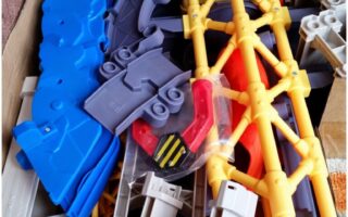 A cardboard box filled with colourful plastic toy track pieces, including blue curved tracks, yellow and grey support structures, and a red and black component, all jumbled together and ready for assembly.