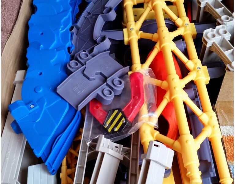 A cardboard box filled with colourful plastic toy track pieces, including blue curved tracks, yellow and grey support structures, and a red and black component, all jumbled together and ready for assembly.