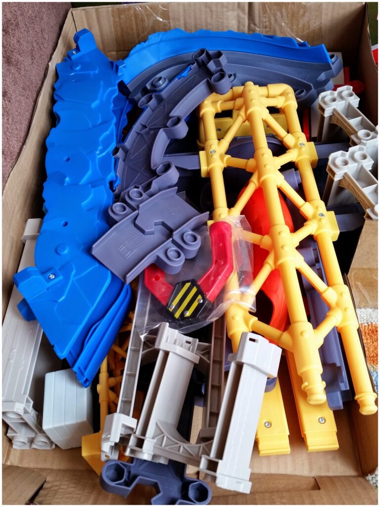 A cardboard box filled with colourful plastic toy train track pieces, including blue, yellow, grey, and red sections, some with support structures and connectors, all stacked haphazardly. The pieces appear to be parts of a buildable train set.
