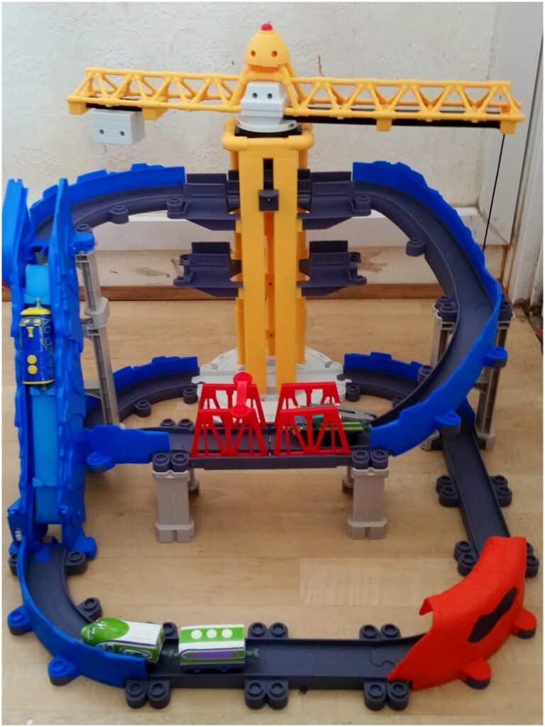 A colourful plastic toy train set with blue and grey tracks, red and grey bridges, a yellow crane, and a red tunnel. Two small green and white toy trains are on the tracks, and a blue lift structure is on the left side.