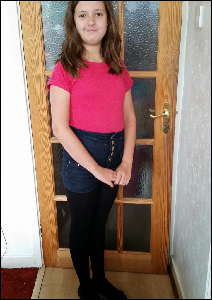 A young girl with long brown hair stands indoors in front of a wooden door with frosted glass panes. She wears a bright pink T-shirt, high-waisted buttoned blue shorts, black tights, and black socks, smiling gently at the camera.