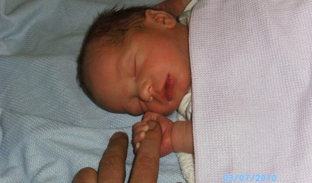 A newborn baby sleeps on a light blue blanket, swaddled in a pale lavender cloth. The baby gently grips an adult’s finger with a tiny hand. The adult’s hand is partially visible. The image is dated 07/03/2010 in blue text at the bottom right.