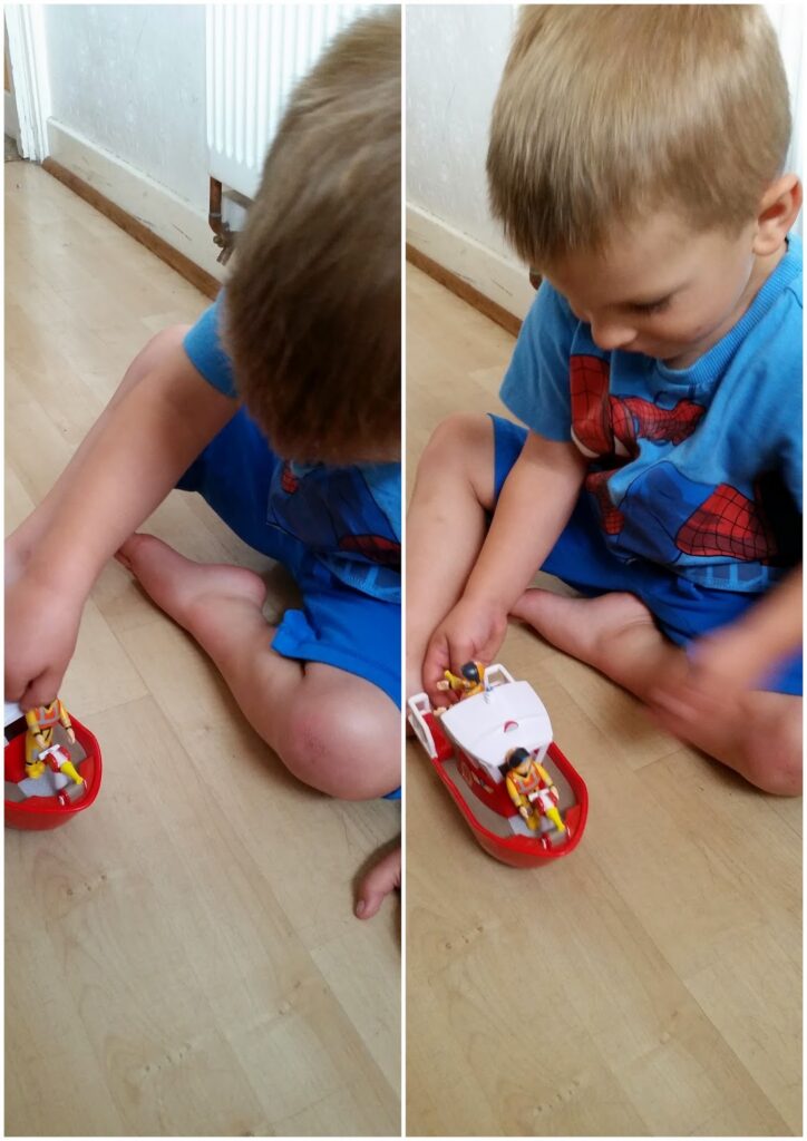 A young child with short blonde hair, wearing a blue T-shirt and shorts, sits on a wooden floor, playing with a small red and white toy boat with figures inside. The child is focused on the boat, using both hands to move and play with it.