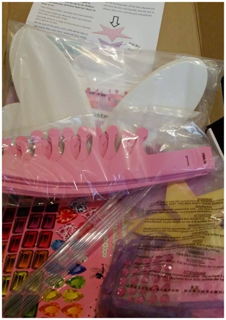 A close-up of a craft kit includes a pink plastic crown with numbered sections, white crown pieces, colourful gem stickers, and instruction sheets. All items are partially wrapped in clear plastic packaging inside a cardboard box.