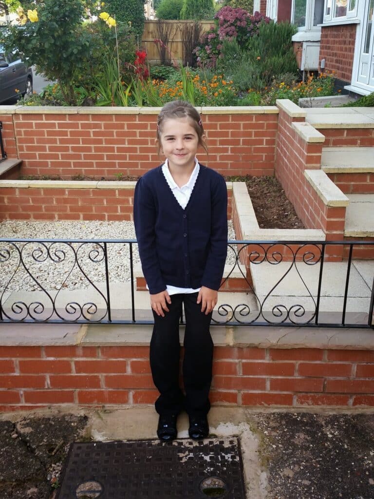 A young girl with brown hair tied back stands on a pavement outside a brick house with tiered gardens. She wears a dark cardigan, white shirt, black trousers, and black shoes, smiling slightly at the camera. Flowers and shrubs are visible behind her.