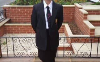 A young girl stands outside in front of a brick house and garden, wearing a black school blazer, white shirt, striped tie, black skirt, tights, and shoes. Her hands are behind her back, and she is smiling at the camera.
