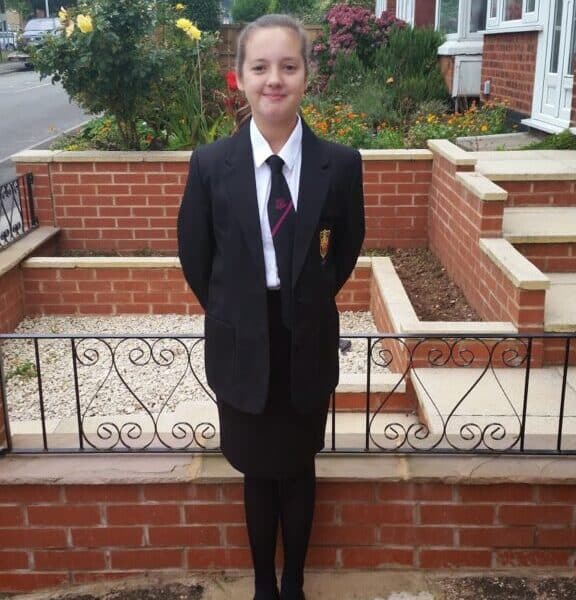 A young girl stands outside in front of a brick house and garden, wearing a black school blazer, white shirt, striped tie, black skirt, tights, and shoes. Her hands are behind her back, and she is smiling at the camera.