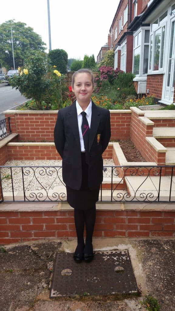 A girl stands outside in front of a brick wall and garden, wearing a black school uniform with a blazer, white shirt, striped tie, knee-length skirt, tights, and black shoes. She is smiling, with her hands behind her back. Houses and greenery are in the background.