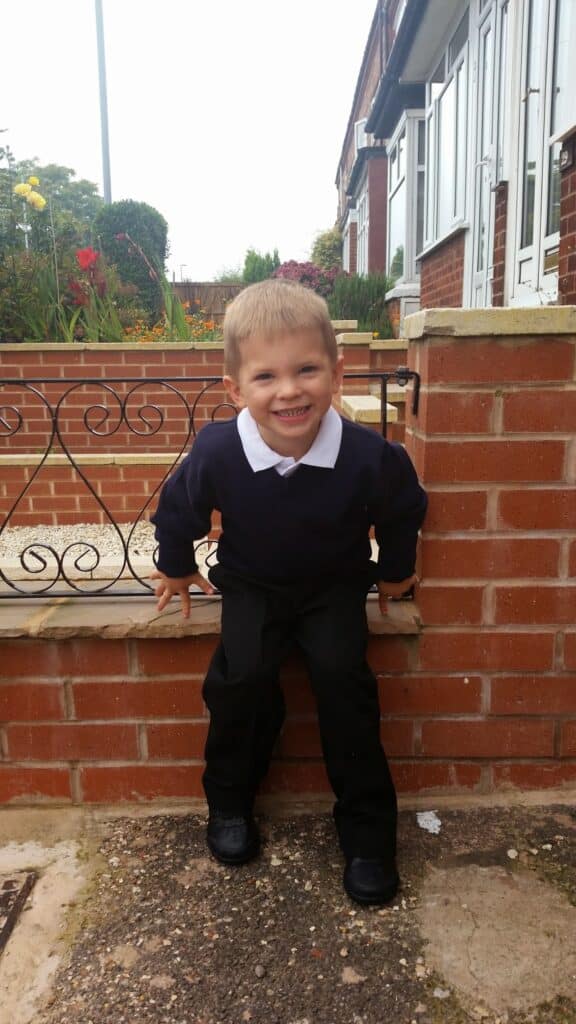 A young boy with short blond hair, wearing a dark jumper, white collared shirt, and black trousers, smiles brightly whilst perched on the edge of a low brick wall in front of a house and garden with flowers.