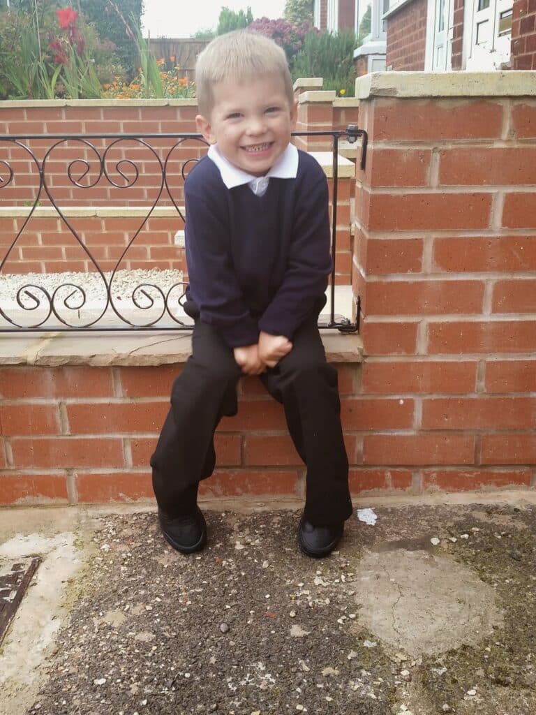 A young boy with short blond hair, wearing a navy blue jumper, white collared shirt, and black trousers, smiles whilst sitting on a brick wall in front of a garden with red flowers and neat brickwork.