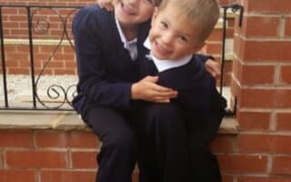 Two young children, a girl and a boy, wearing dark school uniforms with white shirts, sit on a brick ledge outside. The girl hugs the boy, both smiling happily. Behind them are a garden, flowers, and a brick house.
