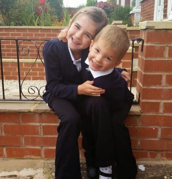Two young children, a girl and a boy, wearing dark school uniforms with white shirts, sit on a brick ledge outside. The girl hugs the boy, both smiling happily. Behind them are a garden, flowers, and a brick house.