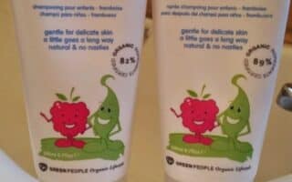 Two tubes labelled “organic children” stand side by side on a sink. The left is shampoo, the right is conditioner, both berry smoothie scent. Each tube features cartoon fruit characters. Swirls of the products are dispensed in front of each tube.