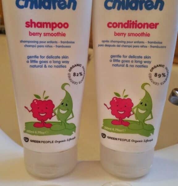 Two tubes labelled “organic children” stand side by side on a sink. The left is shampoo, the right is conditioner, both berry smoothie scent. Each tube features cartoon fruit characters. Swirls of the products are dispensed in front of each tube.