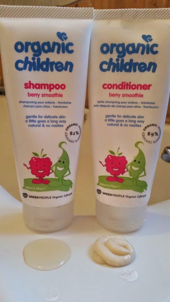 Two tubes labelled "Organic Children" are shown: one is shampoo, the other conditioner, both in "berry smoothie" scent. Each tube features smiling fruit cartoons. In front, a swirl of each product sits on a white surface, with a bathroom sink slightly visible in the background.