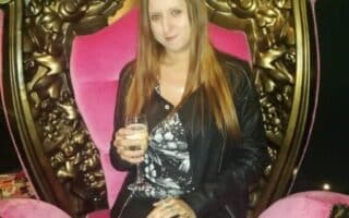 A woman with long straight hair, wearing a black jacket and patterned top, sits on an ornate, gold and pink throne-like chair. She holds a drink in her hand, smiling slightly, with a starry black backdrop behind her.