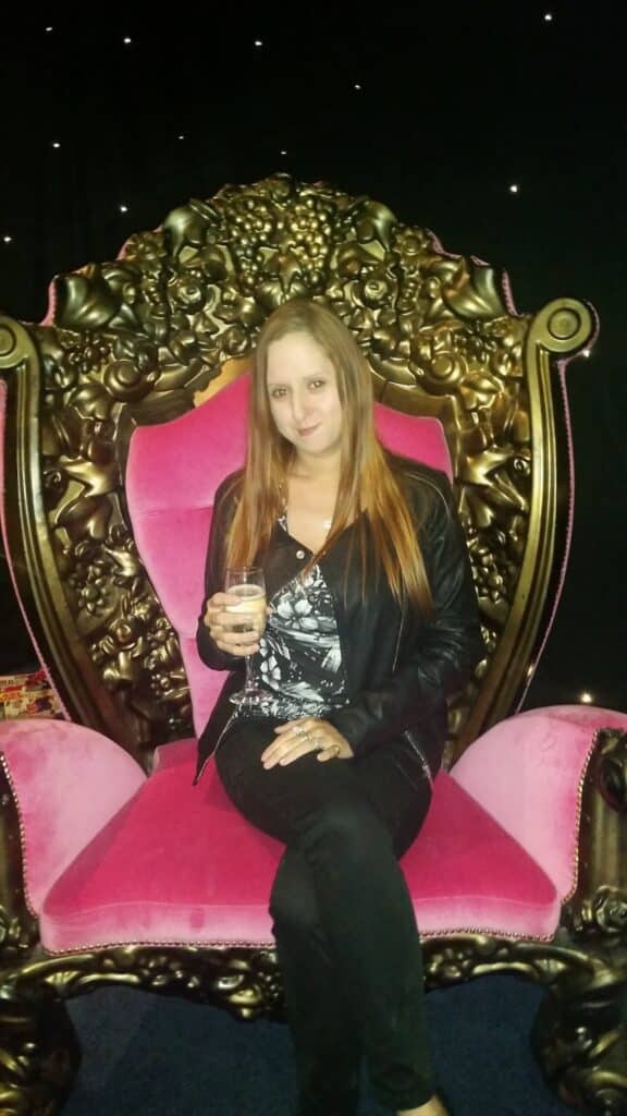 A woman with long straight hair, wearing a black jacket and patterned top, sits on an ornate gold and pink velvet throne chair. She is holding a drink, smiling slightly, with a dark background dotted with small white lights.