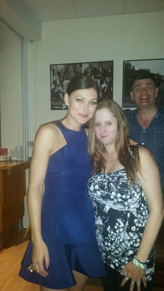 Two women pose together indoors. The woman on the left wears a sleeveless blue dress and has her hair tied back; the woman on the right has long brown hair and a black-and-white floral top. A man in the background photobombs, smiling with an open mouth.