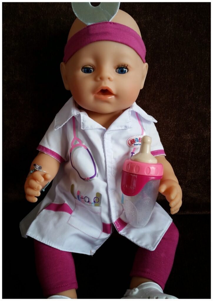 A baby doll dressed as a doctor, wearing a white coat with pink trim, a pink headband with a silver head mirror, and magenta trousers. The doll holds a pink baby bottle and has blue eyes, sitting against a dark background.