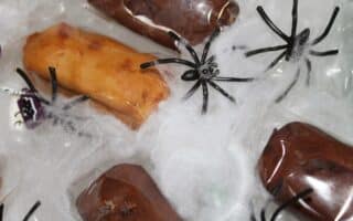 Plastic-wrapped chocolate and golden-brown treats rest on a layer of artificial white cobweb. Several black plastic toy spiders are scattered on top, creating a spooky, Halloween-themed arrangement.