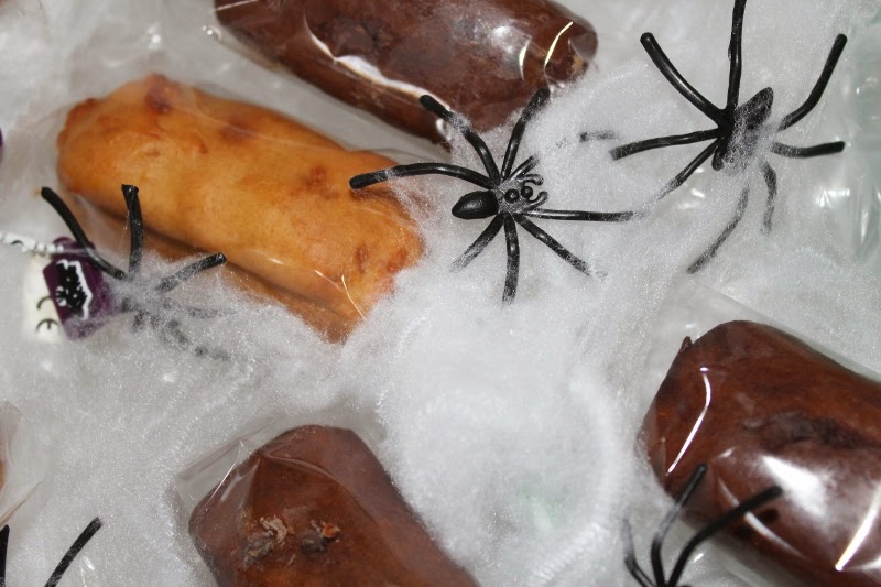 Plastic-wrapped chocolate and golden-brown treats rest on a layer of artificial white cobweb. Several black plastic toy spiders are scattered on top, creating a spooky, Halloween-themed arrangement.