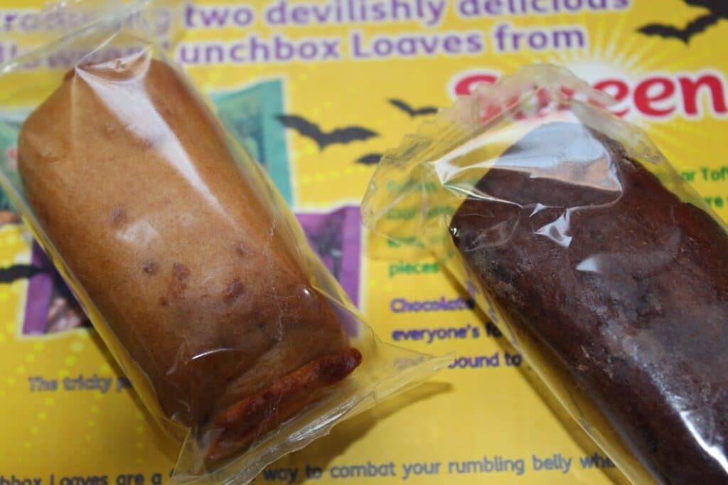 Two small loaf cakes, one light brown and one dark brown, are individually wrapped in clear plastic. They are placed on a colourful background with yellow and purple text, featuring images of black bats and partial wording related to lunchbox loaves.