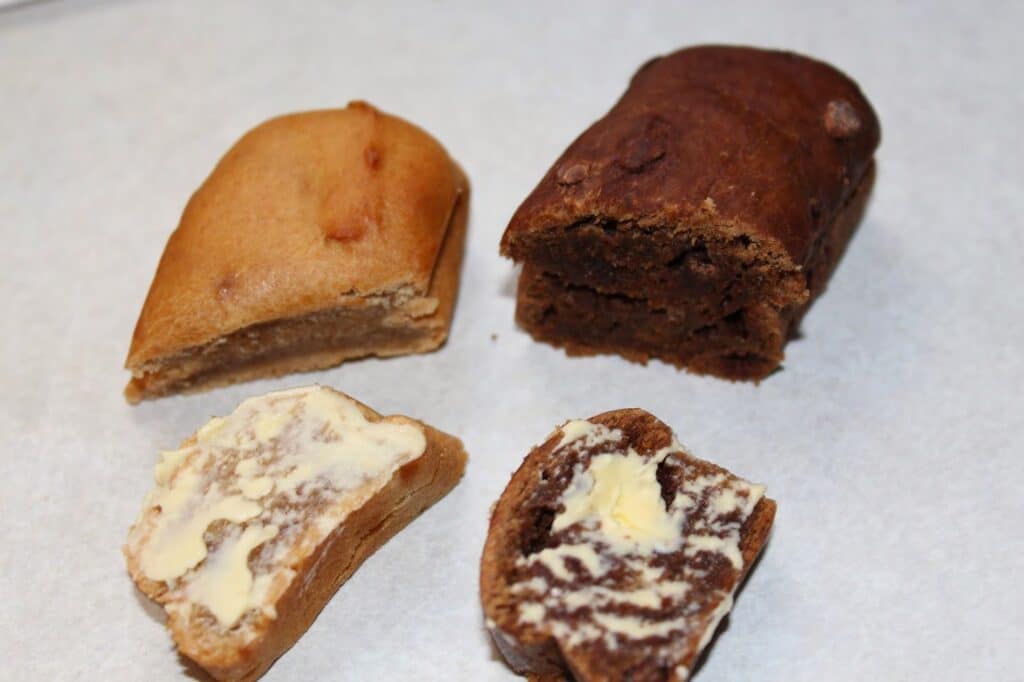 Two small loaves of bread, one light and one dark, sit on a white surface. Each is sliced; the slices in front are spread with butter. The bread appears soft and dense, and the image highlights the contrast between the two types.