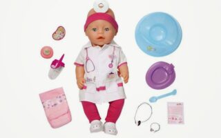 A baby doll dressed as a doctor with a white coat, pink headband, and pink trousers is surrounded by accessories: a nappy, dummy, bottle, stethoscope, syringe, hospital bracelet, ID tag, and a blue feeding set with a plate, spoon, and cup.