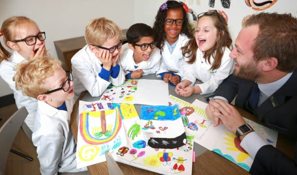 Six young children wearing white lab coats and spectacles sit around a table with a man in a suit. They are smiling and laughing while looking at colourful drawings spread out on the table, suggesting a fun, creative classroom or science activity.