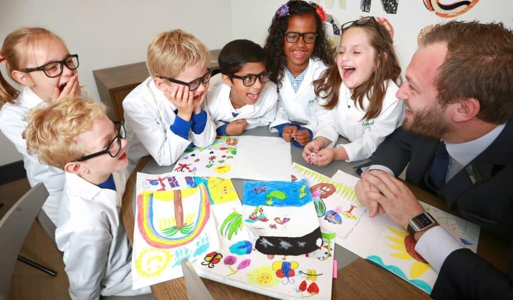 Six young children wearing white lab coats and spectacles sit around a table with a man in a suit. They are smiling and laughing while looking at colourful drawings spread out on the table, suggesting a fun, creative classroom or science activity.
