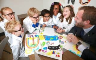 Six young children wearing white lab coats and spectacles sit around a table with a man in a suit. They are smiling and laughing while looking at colourful drawings spread out on the table, suggesting a fun, creative classroom or science activity.