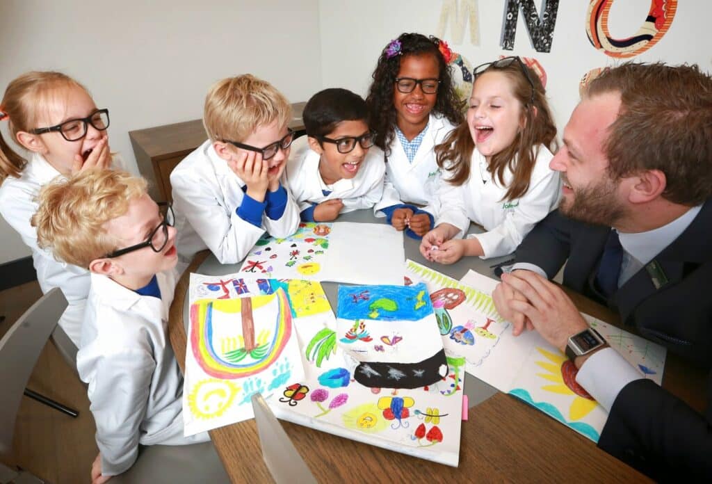 Six children in white lab coats and glasses sit round a table covered with colourful drawings, laughing with an adult man in a suit. The group appears joyful and engaged, suggesting a creative or educational activity in a classroom setting.