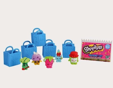 Six blue toy shopping bags and five colourful Shopkins figurines are arranged in front of a pink Shopkins collector’s guide. The figurines resemble various foods with faces, including a strawberry, cupcake, and other cute characters.