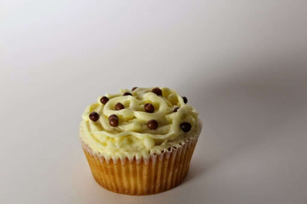 A vanilla cupcake in a paper case is topped with swirls of pale yellow icing and garnished with small round chocolate sweets. The cupcake is set against a plain, light-coloured background with soft lighting and a slight shadow.