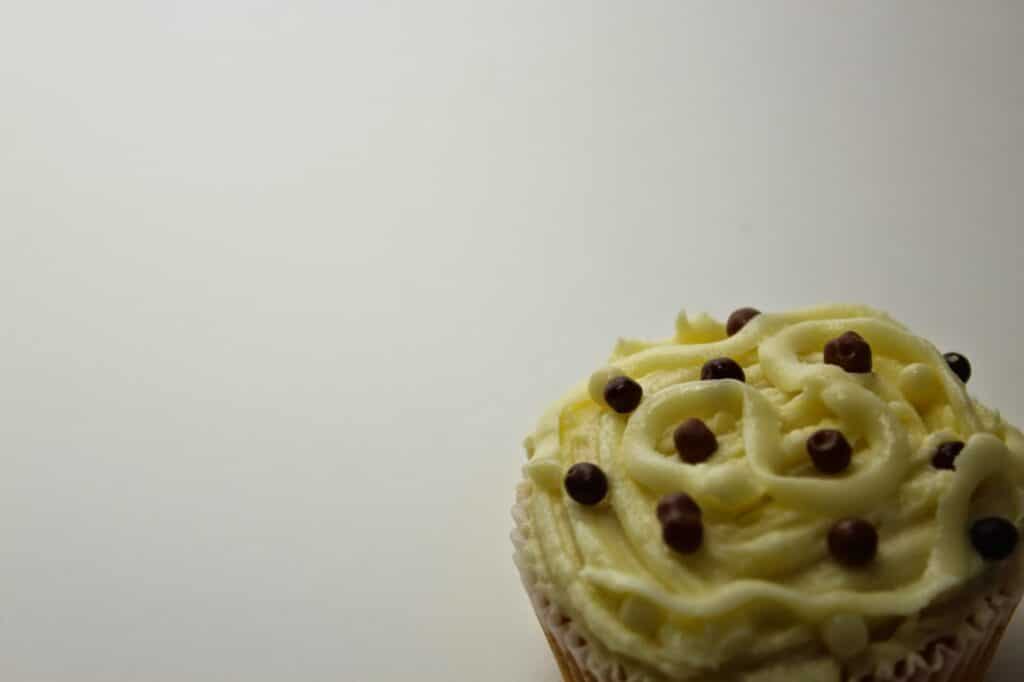 A close-up of a cupcake with creamy white icing, decorated with small round chocolate sweets. The cupcake is positioned in the lower right corner, leaving much of the background empty and white.