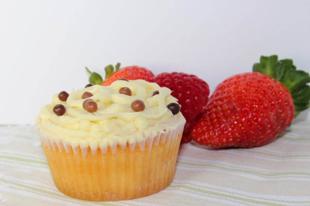 A vanilla cupcake with white icing and brown and white chocolate pearls sits on a striped cloth. Behind it are three ripe strawberries with green tops, against a plain white background.