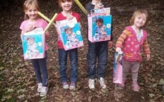 Four young children stand outside on leaf-covered ground, smiling at the camera. Three hold gift bags and toys featuring a cartoon character, while the child on the right holds a striped bag. Trees and a wooden fence are visible in the background.