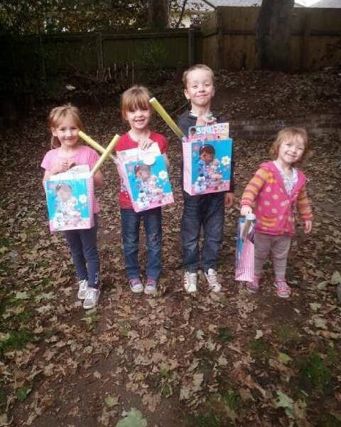 Four young children stand outside on leaf-covered ground, smiling at the camera. Three hold gift bags and toys featuring a cartoon character, while the child on the right holds a striped bag. Trees and a wooden fence are visible in the background.